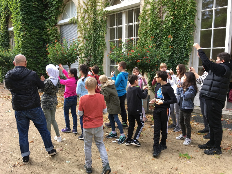 Ein Gruppe Schüler stehen vor einem Gebäude mit Efeubewuchs und schauen dabei alle etwas in der hochgehobenen Wand an.