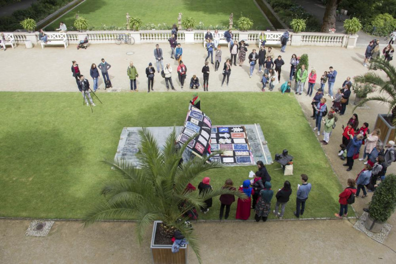 Sicht aus Vogelperspektive: Ein großes, buntes Tuch wird auf einer grünen Wiese aufgefaltet, drum herum stehen Menschen und ein Baum