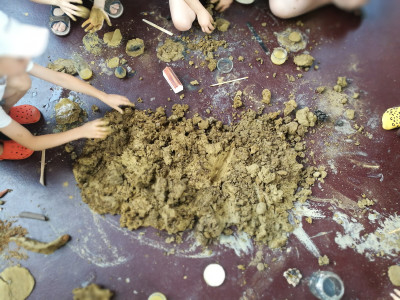 Verschiedene Kinderhände bewegen gelblich braune Erde über einen dunklen Boden, sie formen die Erde auch...